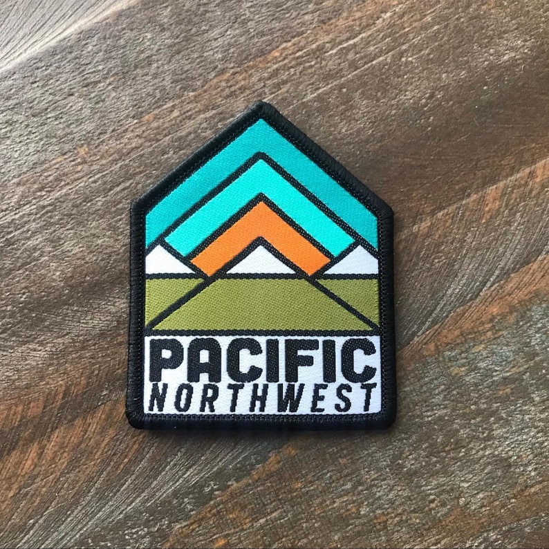 K&ouml;nnte beinhalten: Ein schwarzer und wei&szlig;er Patch mit einer Grafik einer Bergkette mit einem t&uuml;rkisfarbenen Himmel und einer orangefarbenen Sonne. Der Text "PACIFIC NORTHWEST" ist in Schwarz unter der Grafik gedruckt.
