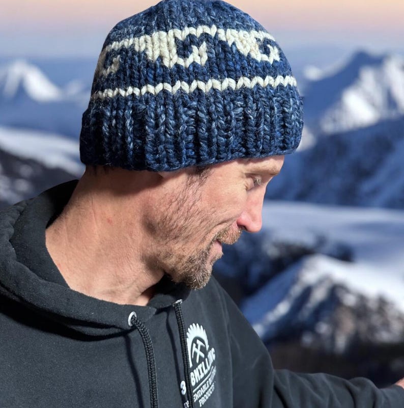 Op de afbeelding: Een man draagt ​​een blauwe en witte gebreide beanie met een geometrisch patroon. De beanie heeft een witte band met een herhalend patroon van kleine witte vierkanten. De man draagt ​​een zwarte hoodie met een wit logo dat "By Billing" zegt en een afbeelding van een zaag en een bijl.