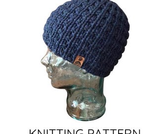 May include: A navy blue knitted beanie hat with a textured pattern. The hat is displayed on a clear mannequin head. A small brown leather tag is sewn on the side. The text "KNITTING PATTERN" is below the hat.