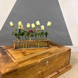 May include: A wooden test tube vase with white roses and small pink flowers. The vase is on a wooden chest with a brass keyhole. The background is a grey and white wall.