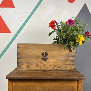 May include: A wooden crate with the number "2" painted on it. The crate is sitting on a wooden table. There are flowers in a vase behind the crate.