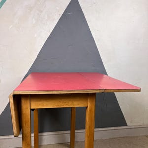 May include: A small, wooden drop-leaf table with a bright red tabletop. The table has four legs and a natural wood finish. The tabletop is approximately 76 cm long when fully extended. The table is against a wall with a geometric design.