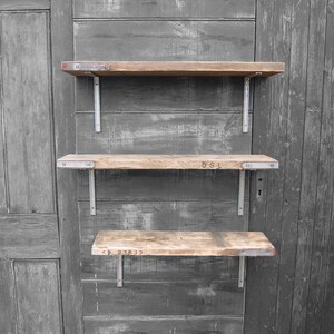 May include: Three wooden shelves with metal brackets mounted on a gray wooden wall. The shelves are made of reclaimed wood and have different lengths. The top shelf has the text "INDUSTRIAL" printed on it. The middle shelf has the text "DSL" printed on it. The bottom shelf has the text "KM 238 32" printed on it.