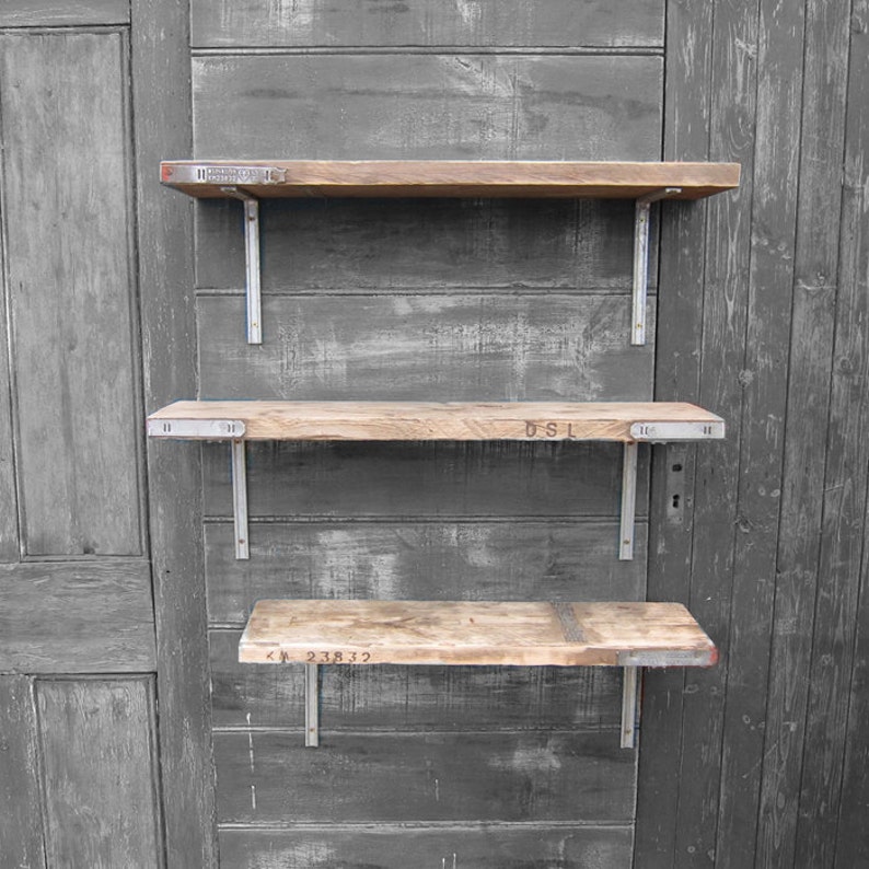 May include: Three wooden shelves with metal brackets mounted on a gray wooden wall. The shelves are made of reclaimed wood and have different lengths. The top shelf has the text "INDUSTRIAL" printed on it. The middle shelf has the text "DSL" printed on it. The bottom shelf has the text "KM 238 32" printed on it.