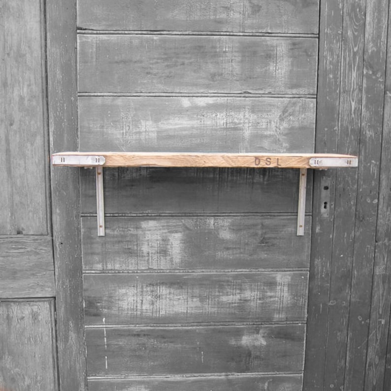 May include: A wooden shelf with metal brackets mounted on a gray wooden wall. The shelf has the letters "DSL" stamped on the surface.