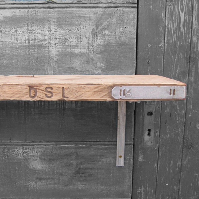 May include: A wooden shelf with a rustic look, featuring the letters "DSL" carved into the front edge. The shelf is supported by two metal brackets, each with a decorative metal plate. The metal plates have the number "16" and a decorative design.