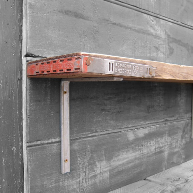 May include: A wooden shelf with a metal bracket attached to a gray wall. The shelf has a metal band with the text "BS1482/08 KH23832" and a heart symbol. The metal bracket is painted silver.