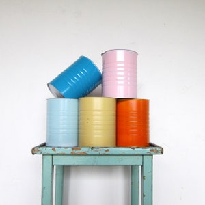 May include: Five colourful metal cans stacked on a light blue wooden table. The cans are light blue, pink, yellow, and orange.