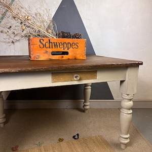 May include: A vintage farmhouse table with a white painted finish and a wooden top. The table has a single drawer and turned legs. A wooden crate with the word "Schweppes" printed on it sits on the table.