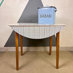 May include: A light blue bread bin with the word "BREAD" in dark letters is on a small, round table. The table has a light wood frame and a checkered pattern top. The table's top is a light blue and white checkered pattern.