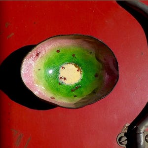 May include: A small, oval-shaped bowl with a colorful enamel design. The bowl features a pink rim, a green center, and a yellow interior. Small brown spots are scattered throughout the design. The bowl is likely used for decorative purposes.