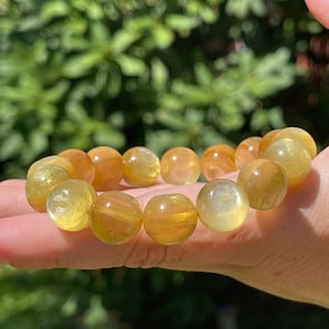 May include: A golden yellow beaded bracelet. The bracelet is made of round, polished beads with varying shades of yellow and orange. The bracelet is displayed against a blurred green background.