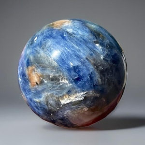 May include: A polished sphere with a swirling pattern of blue, brown, and black hues. The gemstone ball has a smooth, reflective surface, with a hint of red at the base. The sphere is a decorative item.