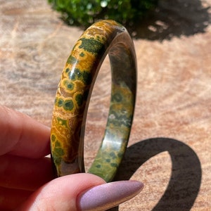 May include: A green and brown patterned bangle bracelet made of polished stone. The bracelet is round and has a smooth surface.