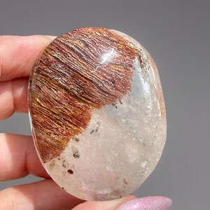 May include: A polished, oval-shaped crystal with a striking combination of colors. The top half features rich, reddish-brown and gold striations, while the bottom half is clear. The crystal is held in a hand, showcasing its natural beauty.