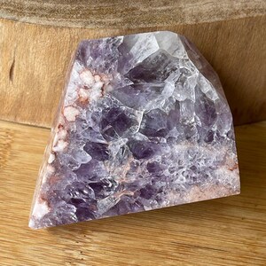 May include: A faceted amethyst crystal with shades of purple, white, and hints of red. The crystal is cut into a triangular shape and has a rough, textured surface. The crystal is displayed on a wooden surface.