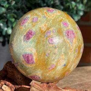 May include: A spherical gemstone with a mottled green and gold surface, speckled with vibrant pink inclusions. The polished stone is displayed on a wooden base, showcasing its natural beauty. This decorative sphere is a unique home decor item.