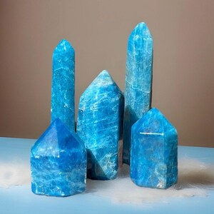 May include: Five blue apatite crystal points of varying heights and shapes. The crystals are a vibrant, translucent blue with white veining. The crystals are arranged on a light blue surface.