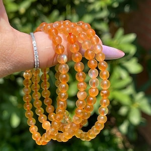 May include: Several strands of orange gemstone bead bracelets are held in a hand. The beads are round and have a translucent quality. A silver ring with small, clear stones is on the hand.