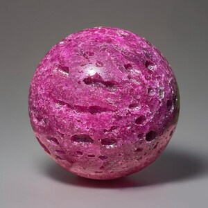 May include: A spherical, textured gemstone in a vibrant magenta hue. The surface is uneven with small, dark holes, giving it a unique, organic appearance. The gemstone is a solid, rounded shape, perfect for display or collection.