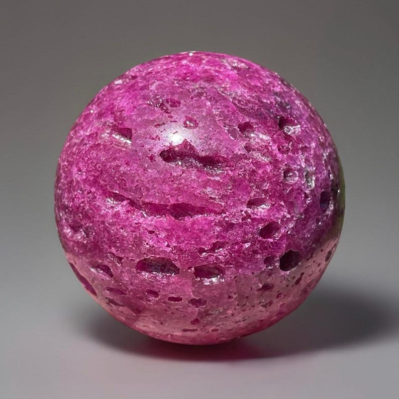 May include: A spherical, textured gemstone in a vibrant magenta hue. The surface is uneven with small, dark holes, giving it a unique, organic appearance. The gemstone is a solid, rounded shape, perfect for display or collection.
