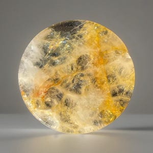 May include: A round, translucent stone with a mix of yellow, gold, and white hues. The stone has a textured, crystalline appearance with dark inclusions. The overall effect is a glowing, natural aesthetic.