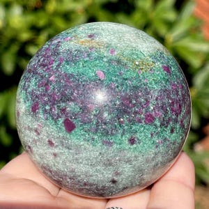 May include: A polished gemstone sphere with a mix of colors. The sphere has a base color of light green with patches of darker green and deep purple. Small flecks of red and purple are scattered throughout the stone.