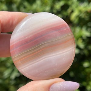 May include: A polished, round stone with bands of white, pink, and reddish-brown hues. The natural stone has a smooth, glossy surface and is held in a hand. The stone's colors and patterns create a unique, organic appearance.