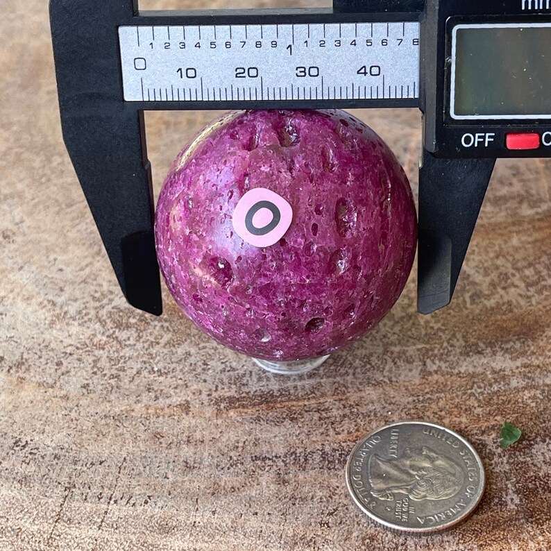 May include: A deep magenta sphere with a textured surface is measured with a digital caliper. The sphere has a small pink sticker with a black circle. A US quarter is in the foreground.
