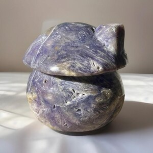 May include: A carved stone box with a lid. The box is a rounded shape with a lid that has a small, raised section. The stone is a mix of purple, gray, and white colors. The box is sitting on a white surface.