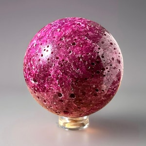 Puede incluir: Objeto decorativo esférico en tonos magenta y rosa oscuro. El orbe tiene una superficie texturizada con numerosos agujeros pequeños y descansa sobre una base circular transparente. El efecto general es el de una gema pulida.