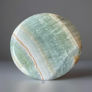 May include: A round, polished stone with a mix of pale green, white, and tan colors. The stone has a smooth surface with visible banding patterns. The overall shape is circular, and the stone appears to be a decorative item.