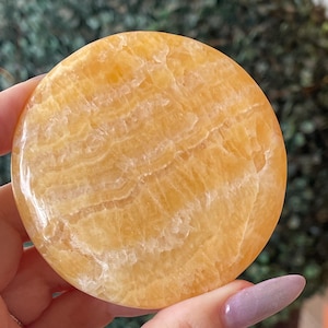 May include: A circular, polished orange calcite stone with a smooth, glossy surface. The stone features natural, translucent bands of varying shades of yellow and orange, creating a layered, marbled appearance throughout the round, flat object.