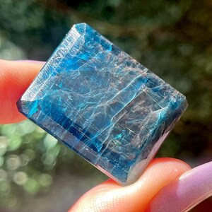 May include: A rectangular, faceted gemstone with a deep blue color and visible internal fractures. The stone has a polished surface with sharp edges and a translucent quality, displaying varying shades of blue throughout its crystalline structure.