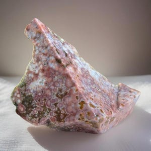 May include: A natural, irregularly shaped stone with a mix of colors. The stone features a base of pink and red hues, with patches of white, green, and brown. The surface has a textured, speckled appearance, and the overall shape is angular.