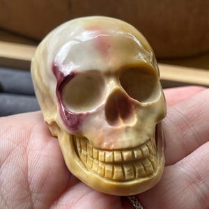 May include: A detailed, carved skull figurine with a cream and tan color scheme, accented by a deep red streak across the left side. The skull features realistic teeth and eye sockets, showcasing intricate craftsmanship.