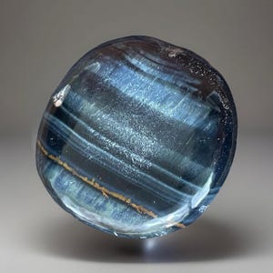 May include: A polished, rounded gemstone with a swirling pattern of deep blue, gray, and gold-brown bands. The stone has a smooth, reflective surface, and the colors create a striking visual effect. The gemstone is a natural mineral specimen.