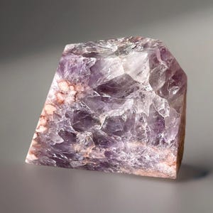 May include: A rough-cut amethyst crystal with a mix of purple, white, and reddish-brown hues. The crystal has a faceted shape and a textured surface, showcasing its natural formation. The gemstone is a decorative item.