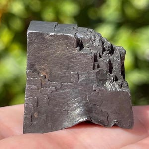 May include: A dark gray, metallic mineral specimen with a cubic, stepped crystal structure. The surface features sharp, geometric edges and a reflective, slightly textured finish, showcasing a blocky, layered formation with distinct, angular facets.