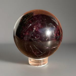 May include: A polished, dark red gemstone sphere, displayed on a clear, round stand. The sphere has a glossy surface with internal inclusions and a deep, rich colour. The stand is transparent, allowing the sphere to be the focal point.