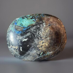 May include: A polished, oval-shaped stone with a mix of colors. The stone features areas of black, brown, teal, and blue. The surface has a glossy finish, and the colors blend together in a natural pattern. The stone is smooth and appears to be a decorative item.