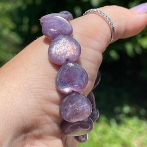 May include: A bracelet featuring heart-shaped beads in shades of purple. The beads have a shimmering, iridescent quality. The bracelet is held in a hand, with a silver ring with small stones on the finger.