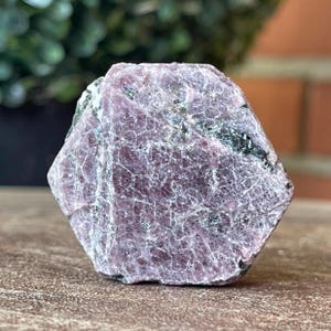 May include: A hexagonal raw crystal specimen featuring a dusty mauve and pink color palette with a textured, cracked surface. The stone displays visible dark grey and black mineral inclusions embedded within its crystalline structure.
