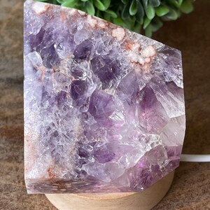 May include: A large, raw amethyst crystal geode, displaying a rich purple hue with white and brown inclusions. The crystal is mounted on a round, light-colored wooden base, creating a natural and earthy aesthetic. The geode is illuminated by a light.