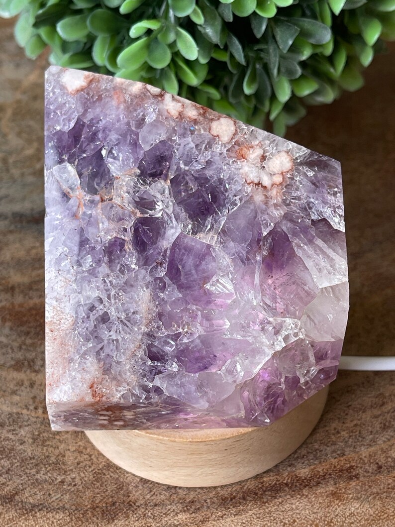 May include: A large, raw amethyst crystal geode, displaying a rich purple hue with white and brown inclusions. The crystal is mounted on a round, light-colored wooden base, creating a natural and earthy aesthetic. The geode is illuminated by a light.