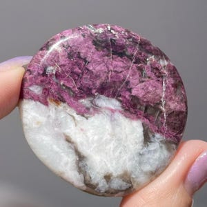 May include: A polished, round gemstone with a striking color contrast. The top half features a rich, textured magenta hue, while the bottom half is a smooth, white with gray veining. The stone is held between two fingers.