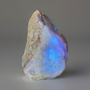 May include: A rough-hewn, natural gemstone with a pale, off-white base color. The stone displays a vibrant, iridescent flash of blue and purple light across its surface. The overall shape is irregular, with a textured, uneven surface.