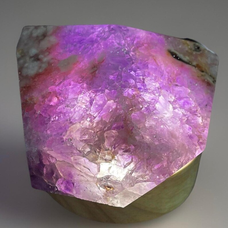 May include: A glowing amethyst crystal geode lamp. The crystal is a vibrant purple with lighter shades and a rough, textured surface. The lamp sits on a wooden base, creating a warm, ambient light.