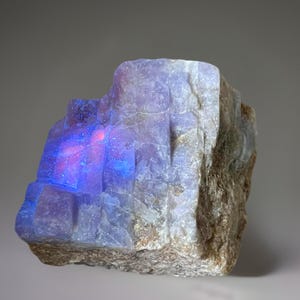 May include: A rough-hewn, natural stone specimen with a striking display of color. The stone features a section with a vibrant, glowing blue and red internal light, contrasting with the muted gray and brown exterior.