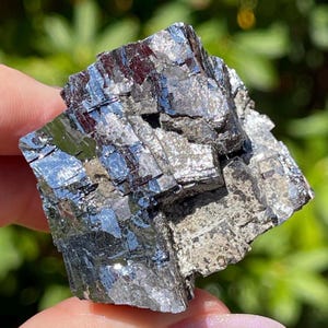 May include: A raw galena mineral specimen featuring a cluster of metallic, silver-gray cubic crystals with sharp, geometric edges and reflective surfaces. The mineral displays a dense, blocky structure with visible striations and deep crevices.